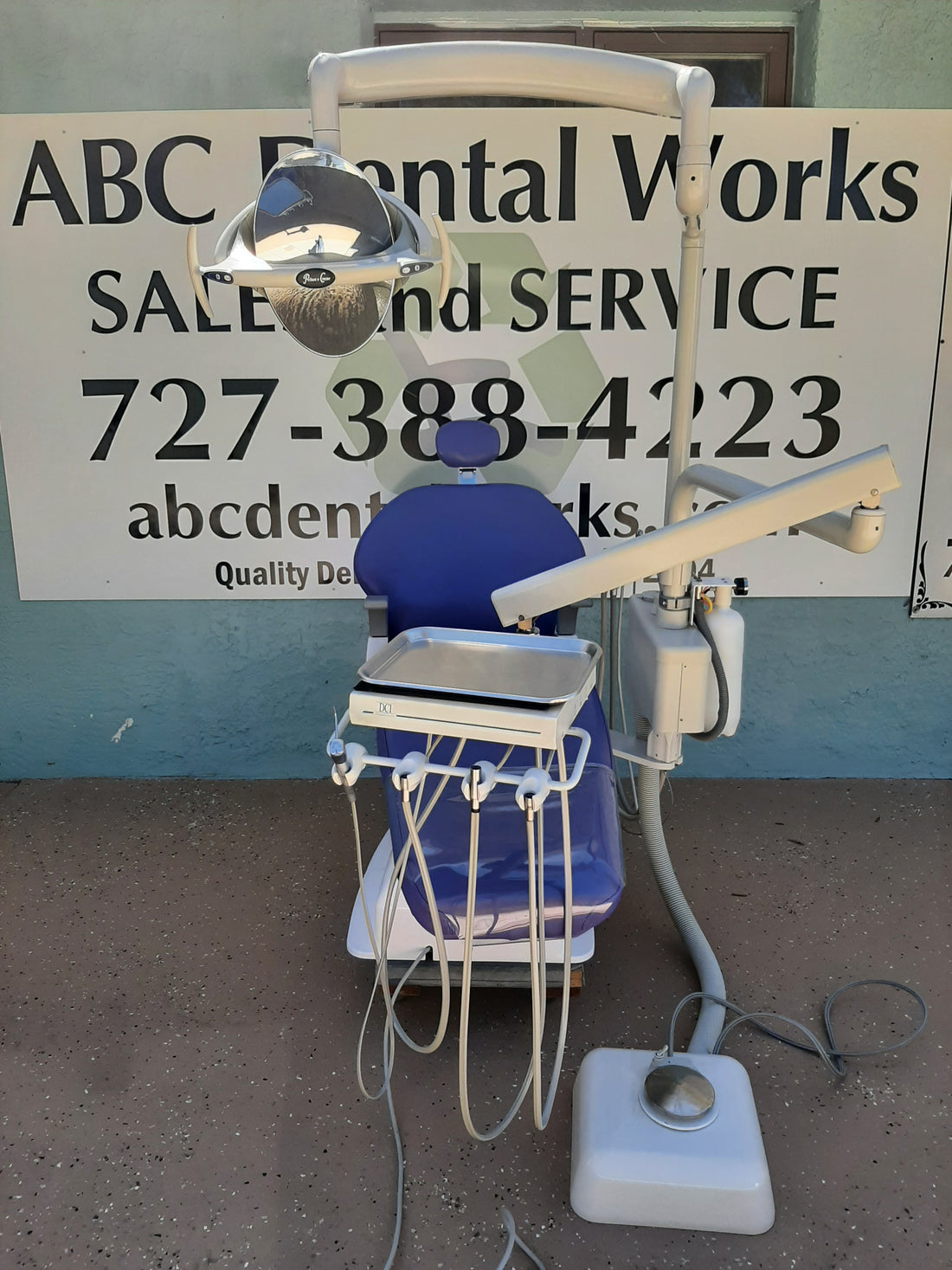 Belmont X-Calibur dental chair front view with blue upholstery, DCI Edge delivery unit, overhead light and foot control on the pavement.