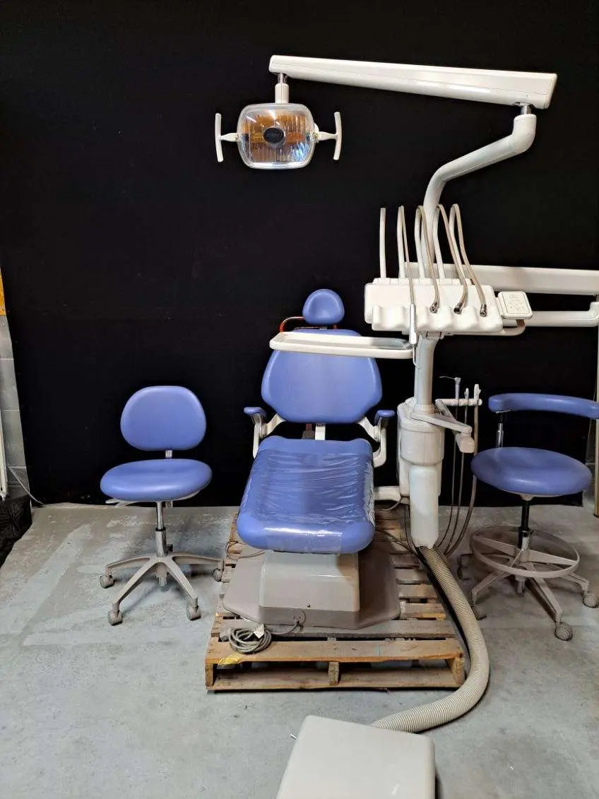 A-dec 1221 package on a pallet: blue upholstered chair, continental delivery with six tubings and A-dec 6300 light overhead; two stools alongside are staging.